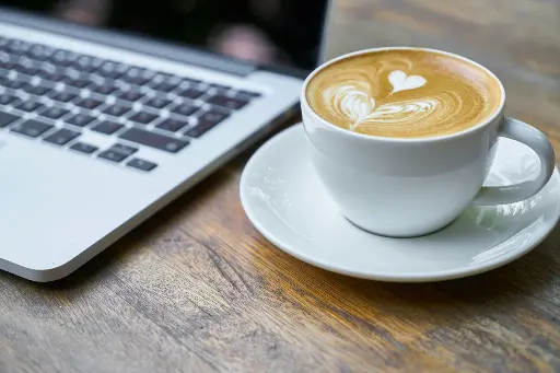 Taza de café latte con arte latte en forma de corazón junto a una laptop sobre una mesa de madera, creando un ambiente acogedor ideal para trabajar, estudiar o disfrutar de café de especialidad.